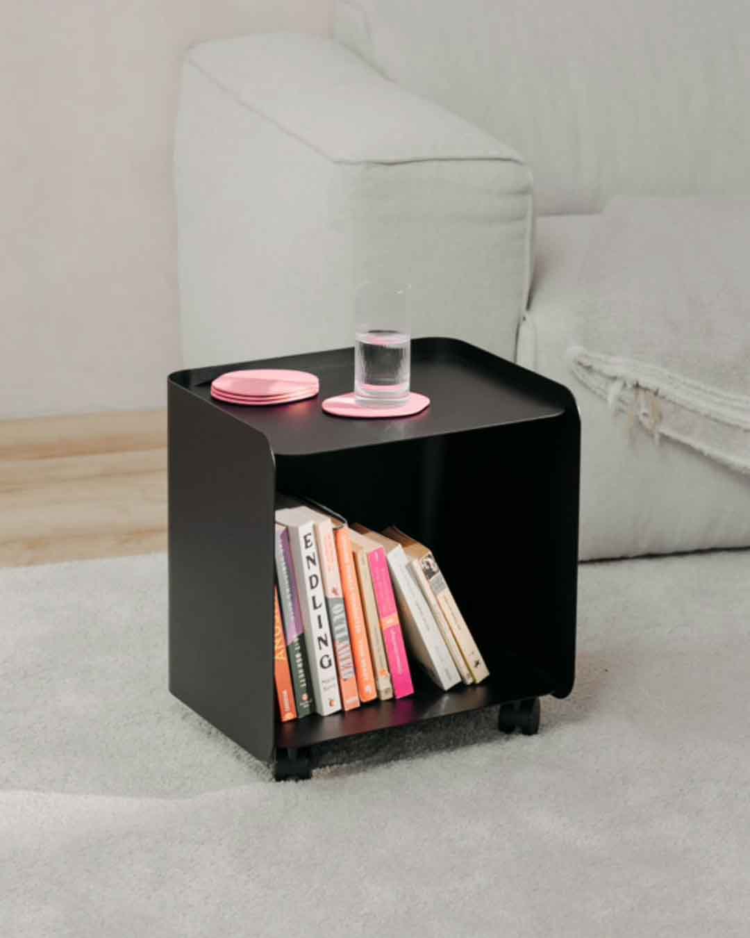 Schwarzer JUNO Rollcontainer in kleiner Ausführung neben einem Sofa mit Büchern und Glas