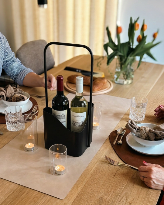 Schwarzer LUNO Flaschenträger aus Stahl mit zwei Weinflaschen auf modern gedecktem Esstisch mit Gläsern, Snacks, Tulpen und warmer Beleuchtung und Personen