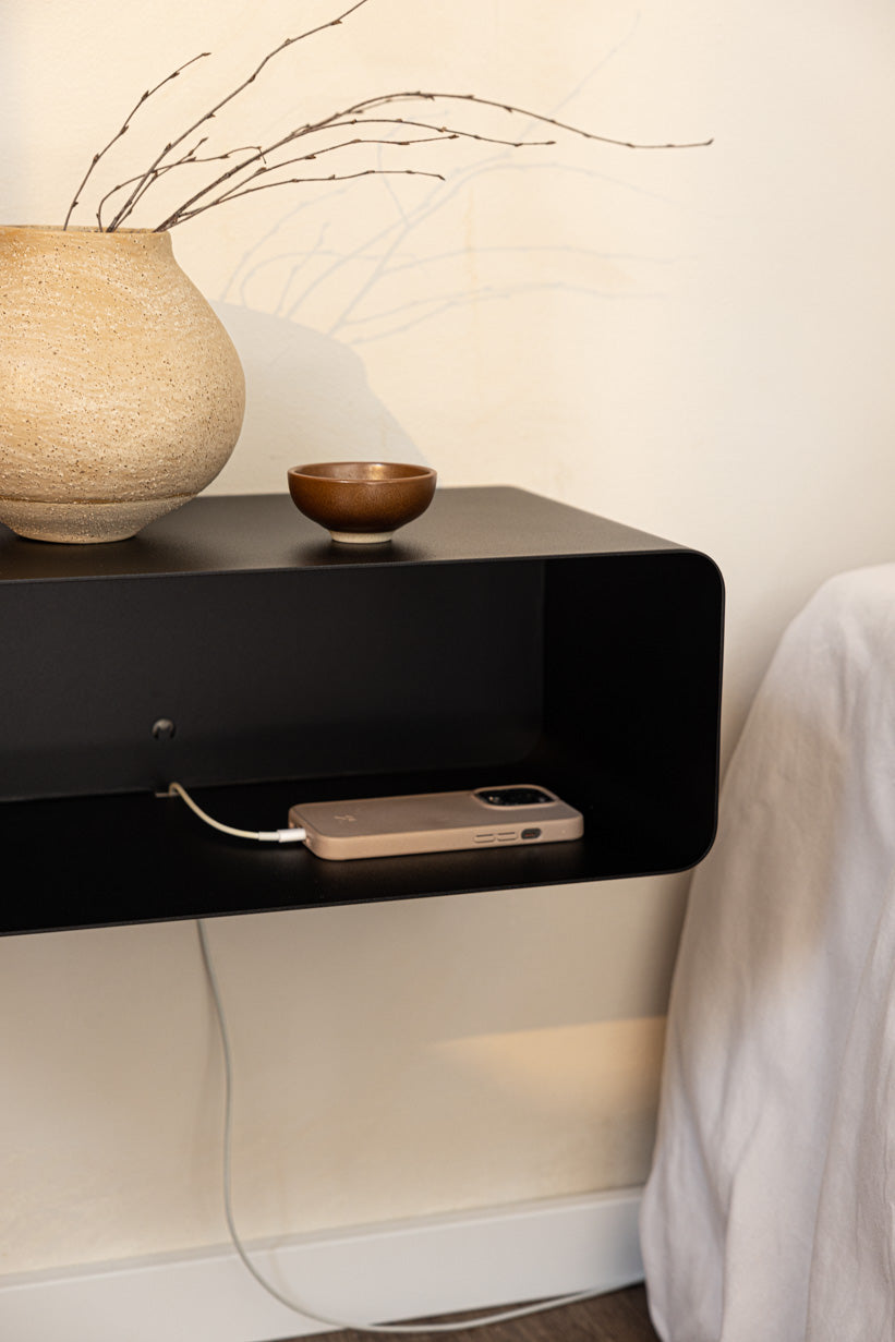 Schwarzer hängender Nachttisch NELIO mit Ablage und Ladekabel neben dem Bett