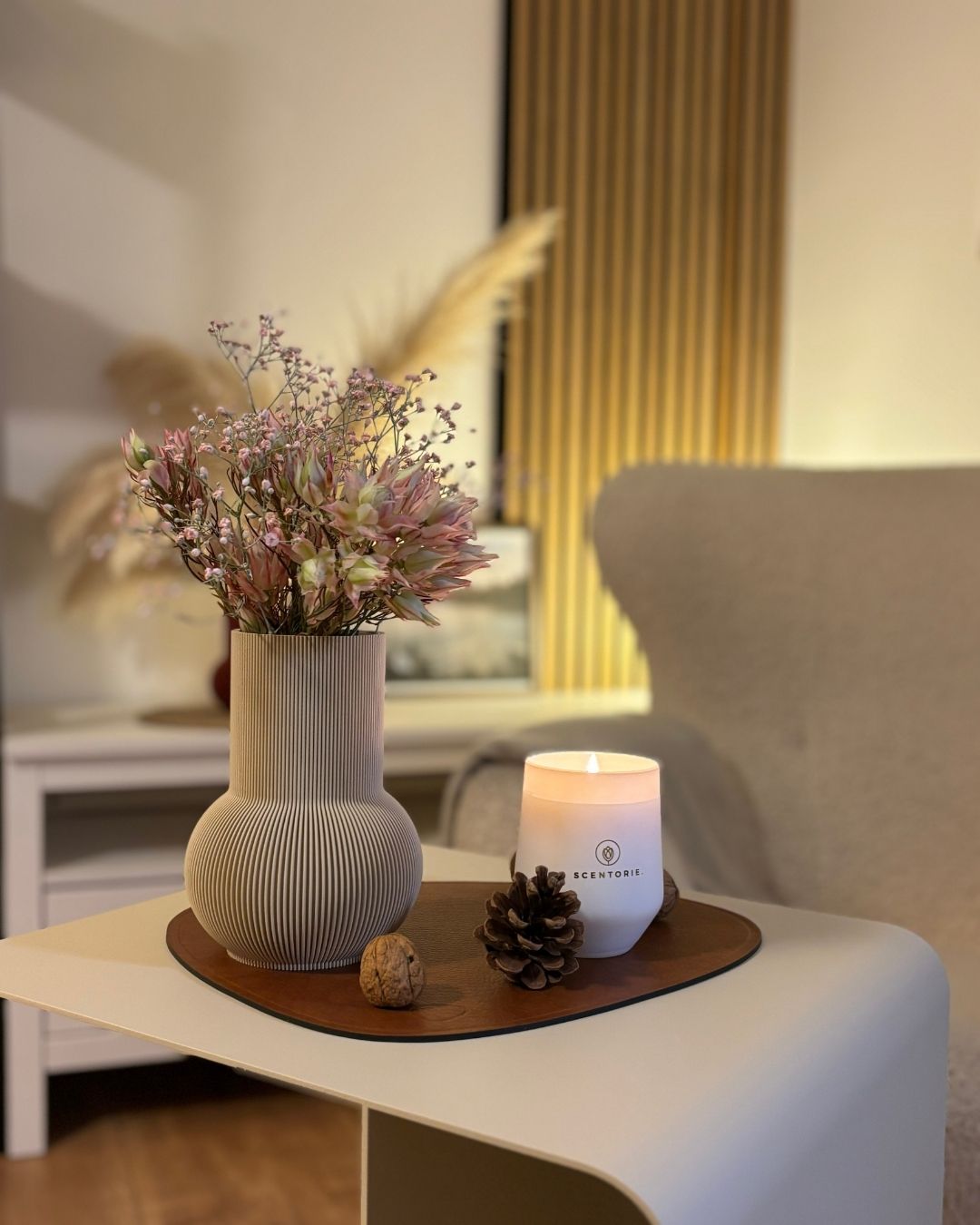 Duftkerze Sunday Morning auf Beistelltisch COSMO und Designunterlage MATS mit Blumen daneben und Docht Feuer an