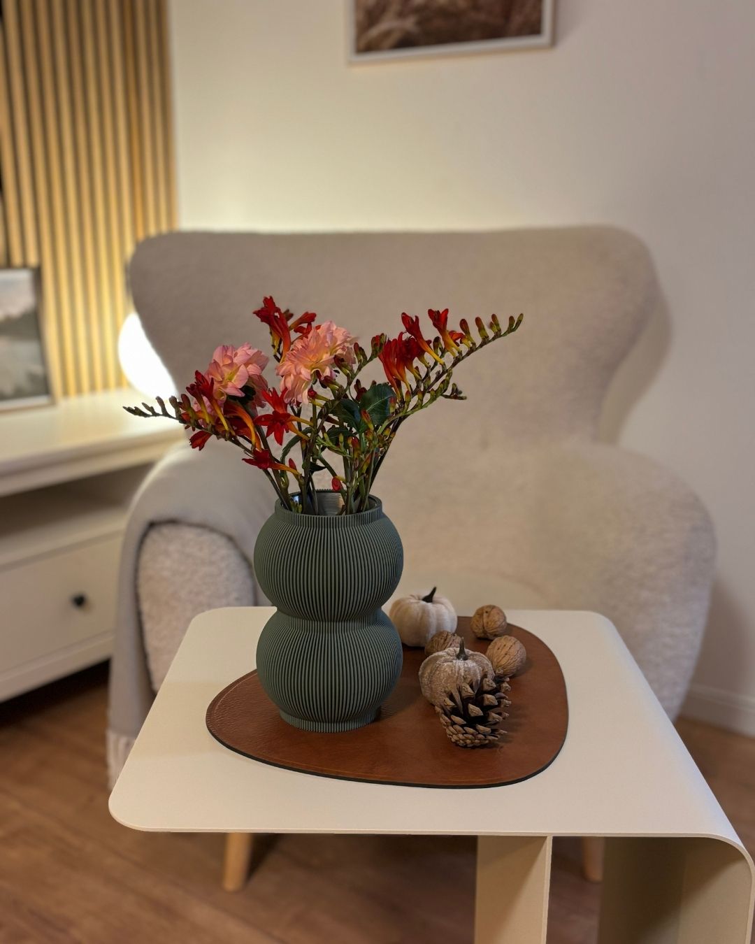 Moosgrüne Designvase SHAPE mit Trockenblumen auf beigem Beistelltisch COSMO mit Sessel im Hintergrund