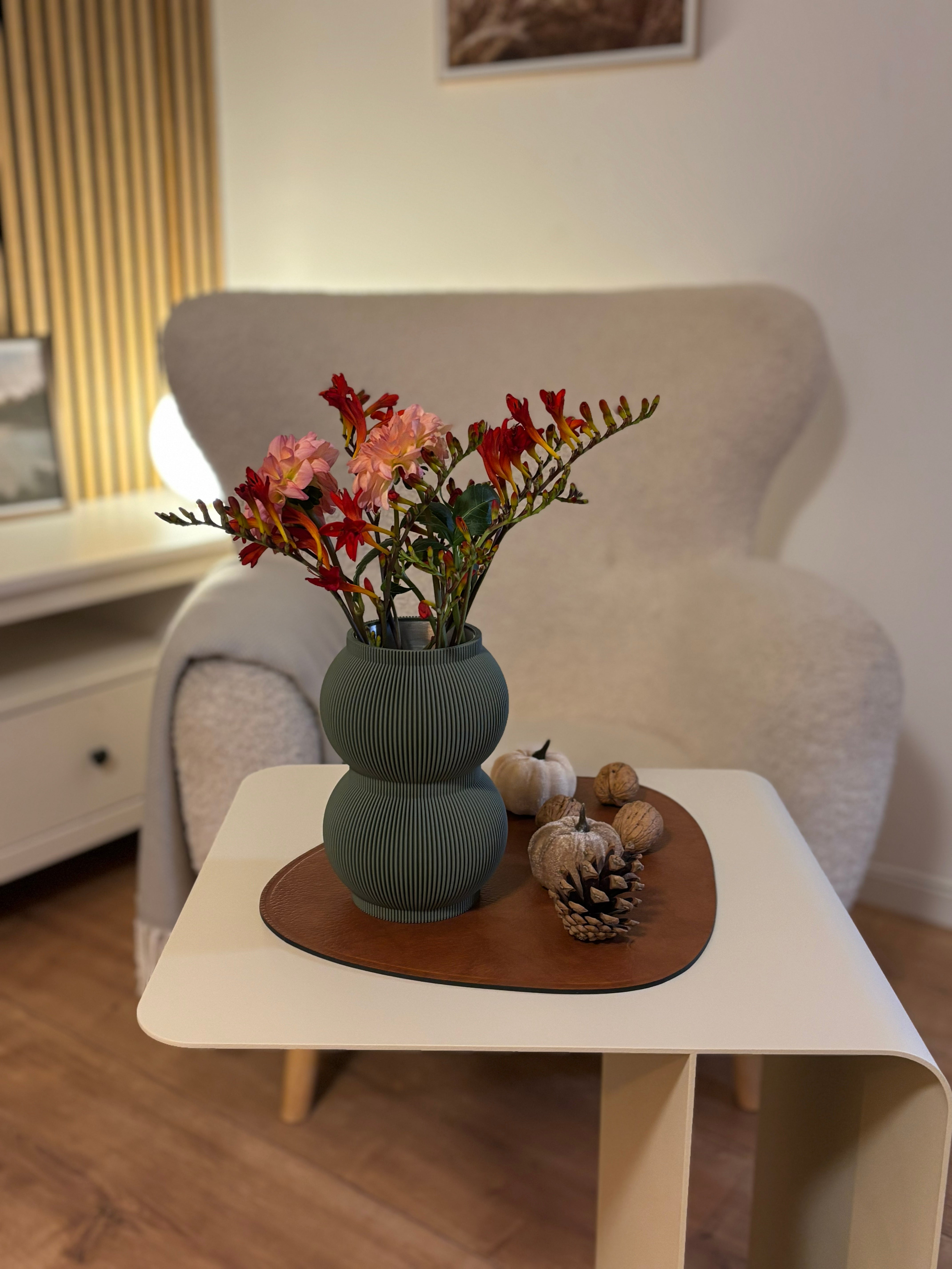 Moosgrüne Designvase SHAPE mit Trockenblumen auf beigem Beistelltisch COSMO mit Sessel im Hintergrund