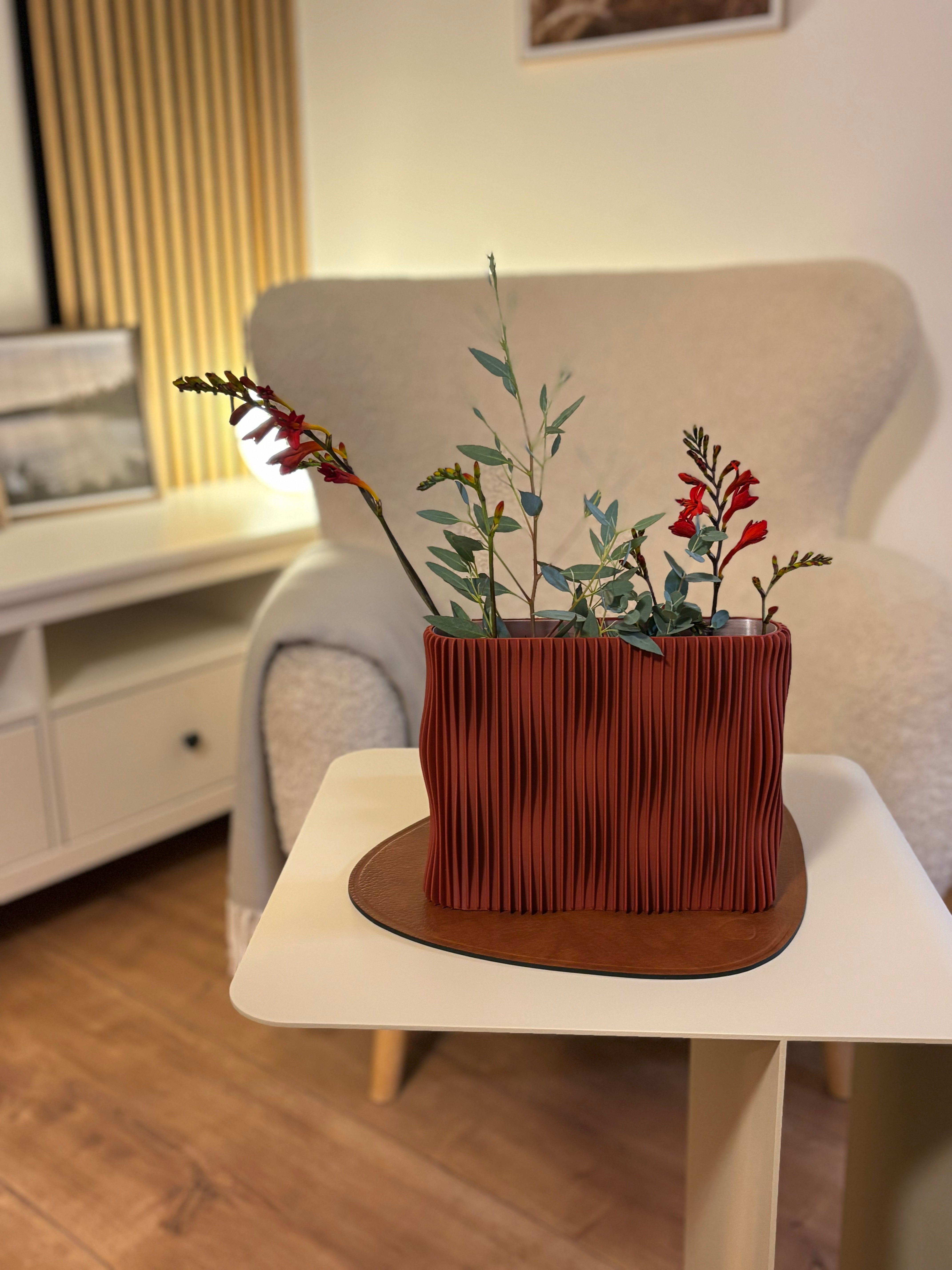 Lehmrote Designvase TWIST mit Trockenblumen auf beigem Beistelltisch COSMO mit Sessel im Hintergrund