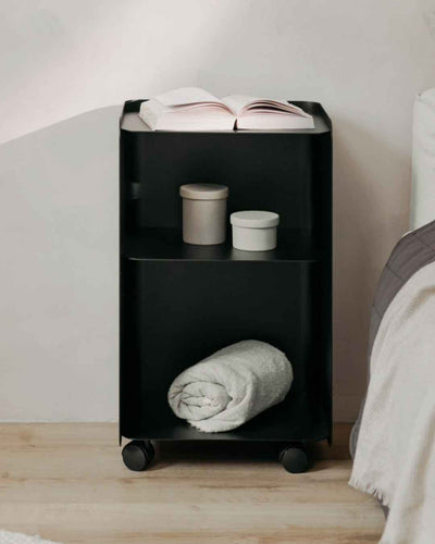 Schwarzer JUNO Rollcontainer in großer Ausführung neben einem Bett mit Buch, Dosen und Decke