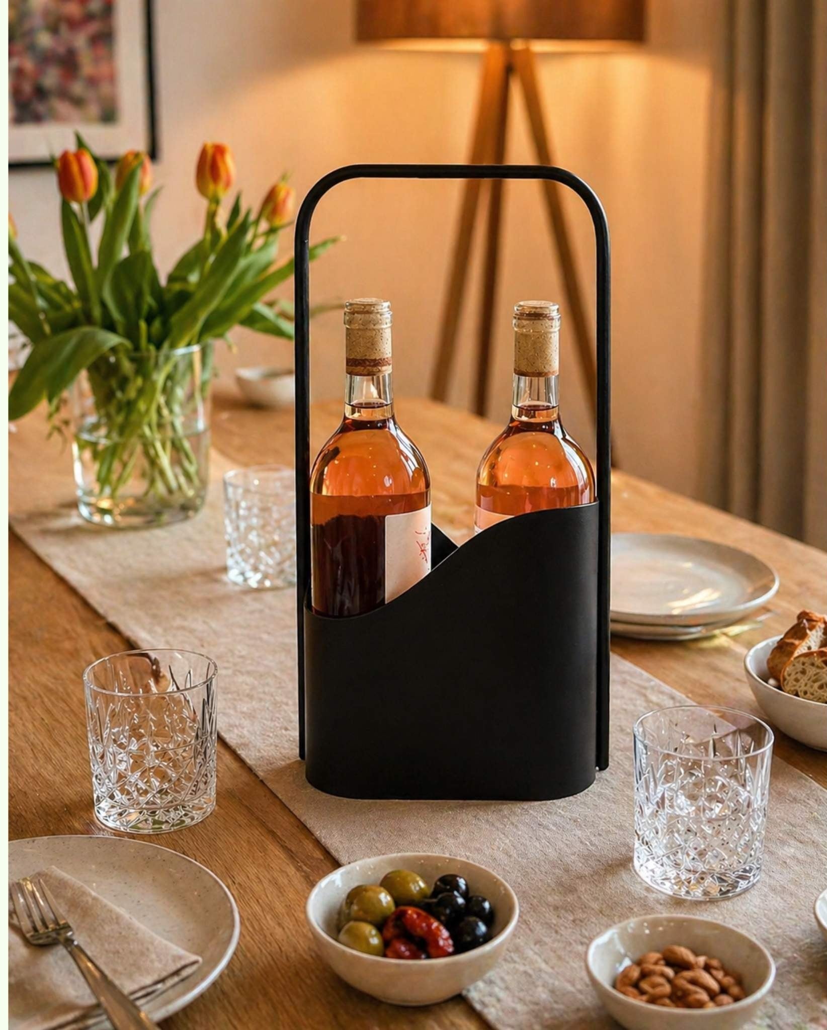 Schwarzer LUNO Flaschenträger aus Stahl mit zwei Weinflaschen auf modern gedecktem Esstisch mit Gläsern, Snacks, Tulpen und warmer Beleuchtung.