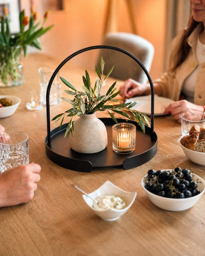 Schwarzes Serviertablett ARCO auf gedecktem Esstisch mit Vase, Kerze, Gläsern und Snacks in gemütlichem Ambiente mit Personen.